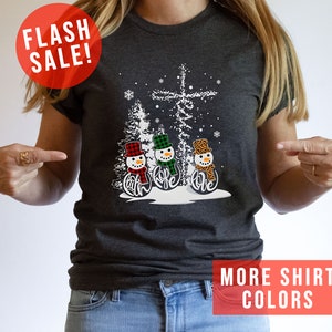 May include: Dark gray t-shirt featuring a winter scene with three snowmen wearing hats and scarves. The snowmen have the words "Faith, Hope, Love" on their bodies. A red circle with "FLASH SALE!" is in the upper left.