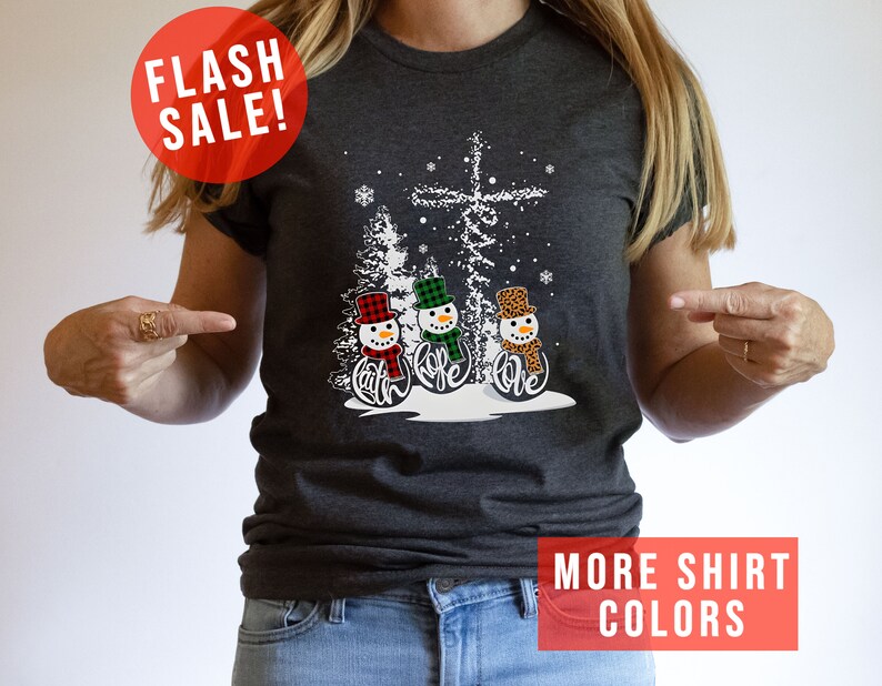 May include: Dark gray t-shirt featuring a winter scene with three snowmen wearing hats and scarves. The snowmen have the words "Faith, Hope, Love" on their bodies. A red circle with "FLASH SALE!" is in the upper left.