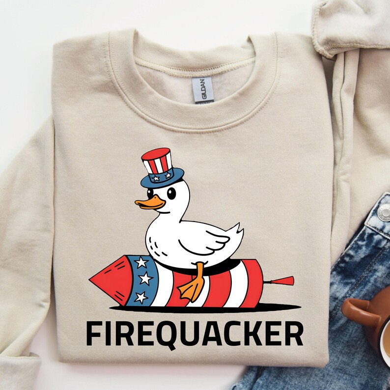 Peut inclure: Sweat-shirt beige avec un canard de dessin anim&eacute; portant un chapeau patriotique et assis sur un feu d'artifice rouge, blanc et bleu. Le mot "FIREQUACKER" est imprim&eacute; en lettres noires en dessous.