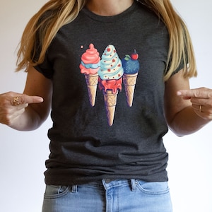 May include: A dark gray t-shirt with a graphic of three ice cream cones. The cones are topped with pink, blue, and purple ice cream and have a cherry, sprinkles, and a swirl design.