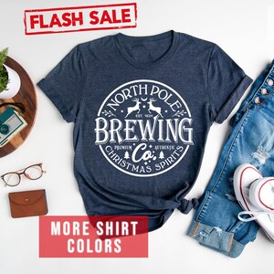 May include: Heather navy t-shirt with a white circular graphic that reads "North Pole Brewing Co. Christmas Spirits." The shirt is displayed with blue jeans and white sneakers. The words "Flash Sale" and "More Shirt Colors" are also visible.