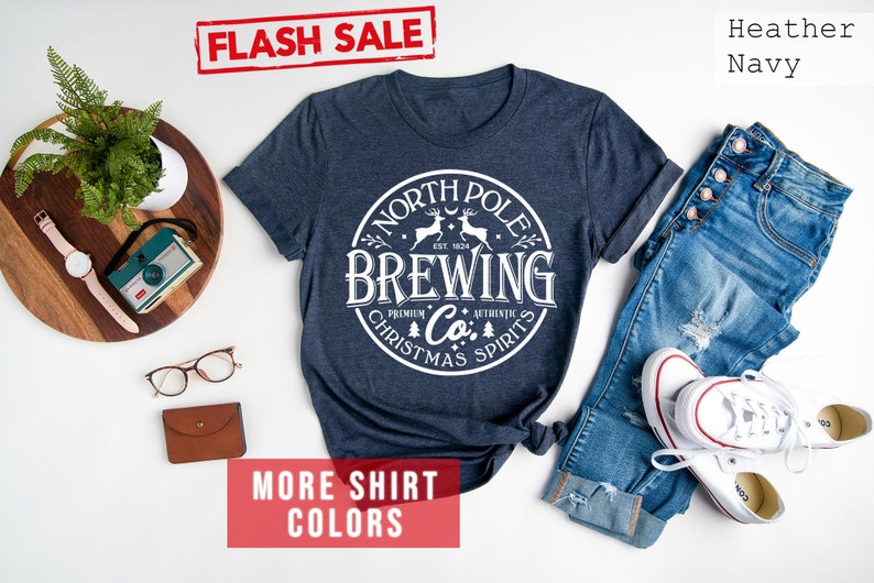 May include: Heather navy t-shirt with a white circular graphic that reads "North Pole Brewing Co. Christmas Spirits." The shirt is displayed with blue jeans and white sneakers. The words "Flash Sale" and "More Shirt Colors" are also visible.