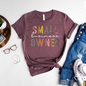 Pode incluir: Uma camiseta marrom-avermelhada com o texto "SMALL business OWNER" em letras coloridas com um padr&atilde;o de leopardo. A camiseta est&aacute; colocada sobre uma superf&iacute;cie branca com um par de cal&ccedil;as jeans, t&ecirc;nis brancos e outros acess&oacute;rios.