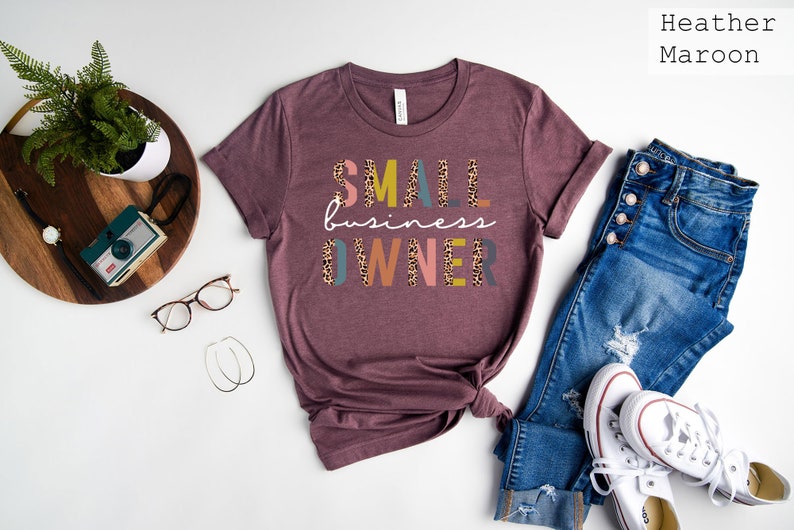 Pode incluir: Uma camiseta marrom-avermelhada com o texto "SMALL business OWNER" em letras coloridas com um padr&atilde;o de leopardo. A camiseta est&aacute; colocada sobre uma superf&iacute;cie branca com um par de cal&ccedil;as jeans, t&ecirc;nis brancos e outros acess&oacute;rios.