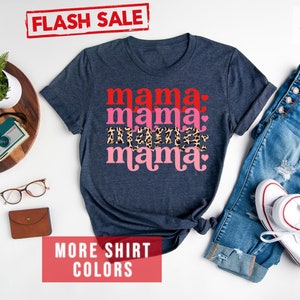 Op de afbeelding: Een marineblauw T-shirt met de tekst "mama mama mama" in rood, roze en luipaardprint. Het shirt ligt plat op een wit oppervlak met een spijkerbroek, een horloge, een bril en een camera.