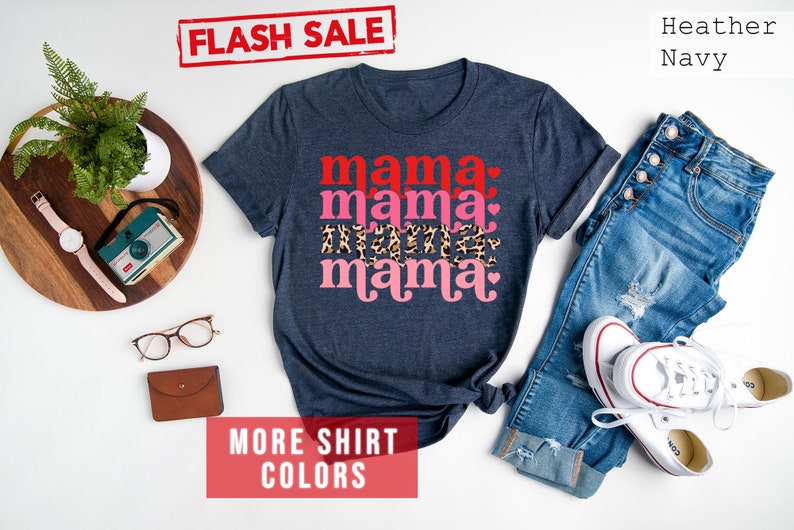 Op de afbeelding: Een marineblauw T-shirt met de tekst "mama mama mama" in rood, roze en luipaardprint. Het shirt ligt plat op een wit oppervlak met een spijkerbroek, een horloge, een bril en een camera.