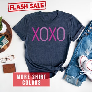 May include: A heather navy blue t-shirt with the word "XOXO" printed in pink on the front. The shirt is laid flat on a white surface with a pair of jeans and white trainers. The text "FLASH SALE" and "MORE SHIRT COLORS" are printed in red on white rectangles.