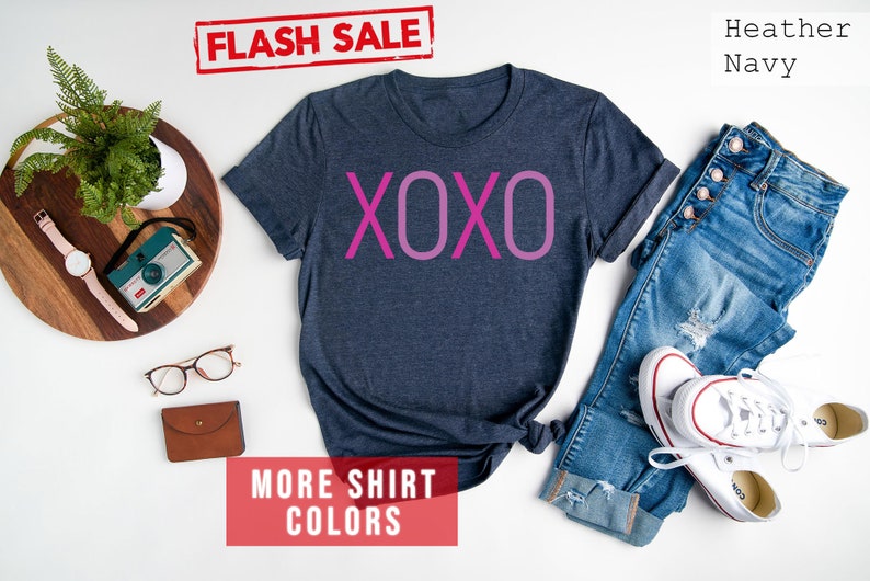 May include: A heather navy blue t-shirt with the word "XOXO" printed in pink on the front. The shirt is laid flat on a white surface with a pair of jeans and white trainers. The text "FLASH SALE" and "MORE SHIRT COLORS" are printed in red on white rectangles.