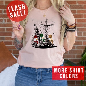 May include: A light pink t-shirt featuring a graphic of three snowmen with the words "Faith, Hope, Love" in cursive. The snowmen wear hats and scarves in plaid and leopard print. A cross and Christmas trees are in the background. The words "FLASH SALE!" and "MORE SHIRT COLORS" are also visible.