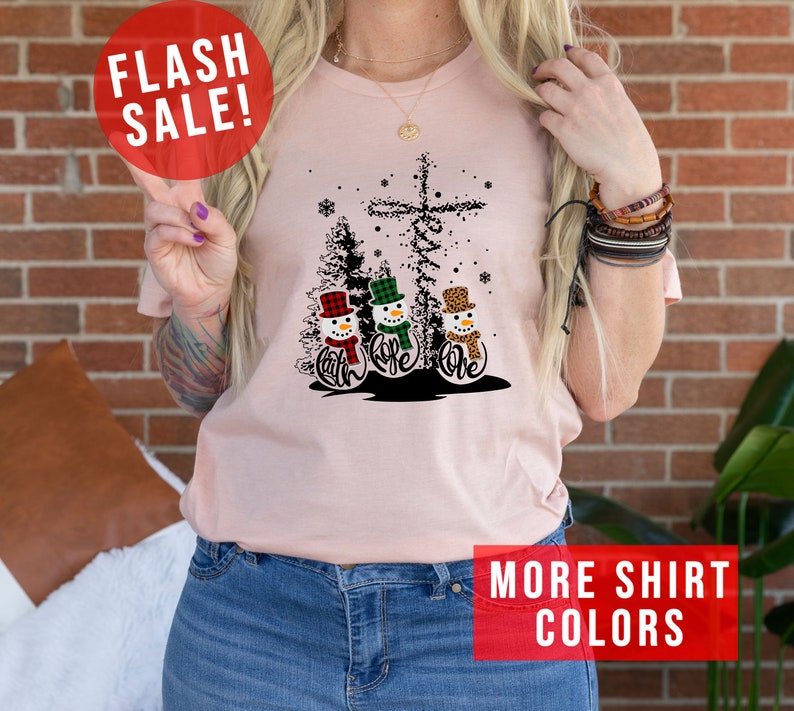 May include: A light pink t-shirt featuring a graphic of three snowmen with the words "Faith, Hope, Love" in cursive. The snowmen wear hats and scarves in plaid and leopard print. A cross and Christmas trees are in the background. The words "FLASH SALE!" and "MORE SHIRT COLORS" are also visible.