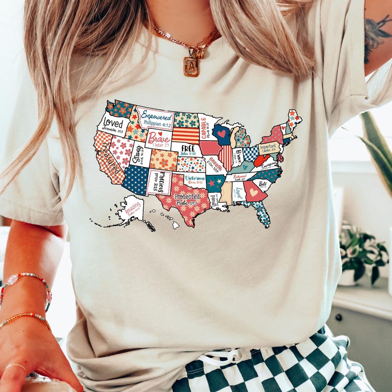 May include: A beige t-shirt with a graphic of the United States of America. Each state is a different color and pattern with words like "Loved", "Brave", "Free", "Protected", and "Enough" written on them.