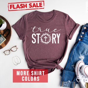 May include: A heather maroon t-shirt with the text "true STORY" in white lettering. A white cross is inside a crown of thorns. The shirt is laid flat on a white surface with a pair of jeans, white sneakers, and other accessories.