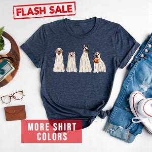 Op de afbeelding: Een heather navy t-shirt met korte mouwen en een afbeelding van vier honden in witte spookkostuums, waarbij &eacute;&eacute;n hond een kleine oranje pompoen vasthoudt. Het shirt wordt getoond naast een blauwe spijkerbroek, witte sneakers en diverse accessoires.