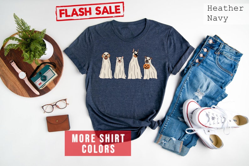 Op de afbeelding: Een heather navy t-shirt met korte mouwen en een afbeelding van vier honden in witte spookkostuums, waarbij &eacute;&eacute;n hond een kleine oranje pompoen vasthoudt. Het shirt wordt getoond naast een blauwe spijkerbroek, witte sneakers en diverse accessoires.
