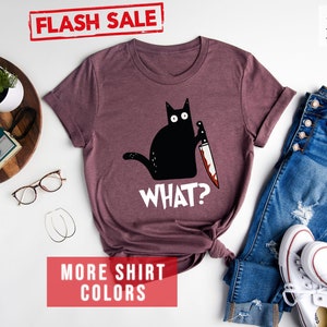 Op de afbeelding: Een heide-marron t-shirt met een zwarte kat die een bloedig mes vasthoudt en de tekst "WHAT?" op de voorkant. Het shirt ligt op een wit oppervlak met een bril, een houten dienblad met een camera en een plant, en een spijkerbroek en witte sneakers.