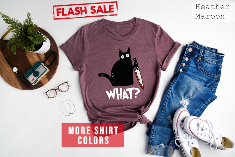 Op de afbeelding: Een heide-marron t-shirt met een zwarte kat die een bloedig mes vasthoudt en de tekst "WHAT?" op de voorkant. Het shirt ligt op een wit oppervlak met een bril, een houten dienblad met een camera en een plant, en een spijkerbroek en witte sneakers.