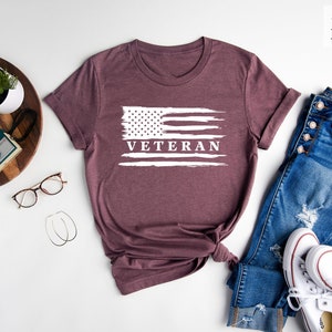 May include: A heather maroon t-shirt with a white distressed American flag and the word "Veteran" printed on it. The shirt is laid flat on a white surface with a pair of jeans, a pair of white sneakers, and a small potted plant.