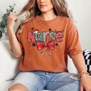 May include: A rust-colored t-shirt with a Christmas-themed graphic that says "Nurse" in colorful letters with a red bow and a stethoscope.
