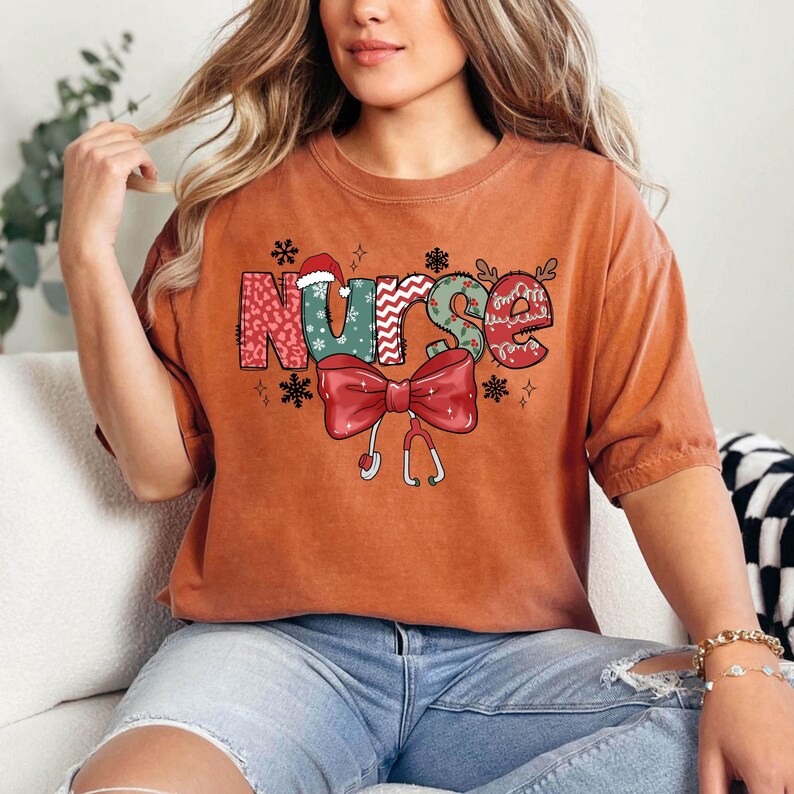 May include: A rust-colored t-shirt with a Christmas-themed graphic that says "Nurse" in colorful letters with a red bow and a stethoscope.