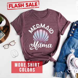 May include: A heather maroon t-shirt with the text "Mermaid Mama" in white. The shirt features a white seashell with a pink and blue ombre design. The text "FLASH SALE" and "MORE SHIRT COLORS" are in red.
