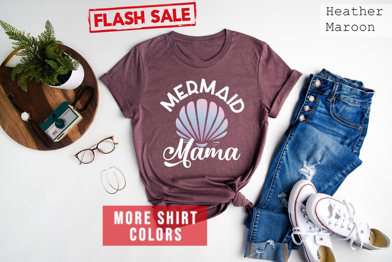 May include: A heather maroon t-shirt with the text "Mermaid Mama" in white. The shirt features a white seashell with a pink and blue ombre design. The text "FLASH SALE" and "MORE SHIRT COLORS" are in red.