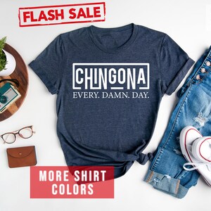 May include: A navy blue t-shirt with white text that reads "CHINGONA EVERY. DAMN. DAY."  The shirt is laid flat on a white surface with a pair of jeans, a pair of glasses, a watch, and a camera.