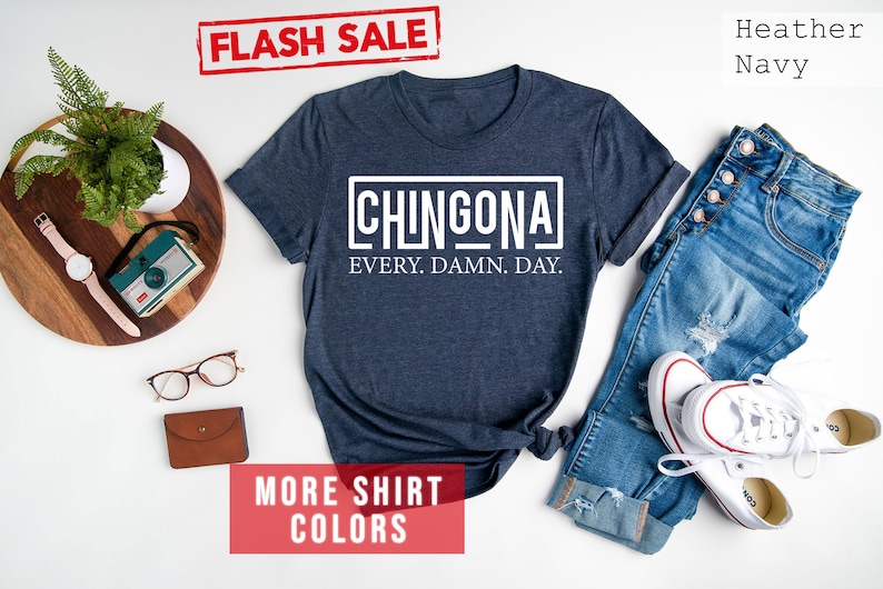 May include: A navy blue t-shirt with white text that reads "CHINGONA EVERY. DAMN. DAY."  The shirt is laid flat on a white surface with a pair of jeans, a pair of glasses, a watch, and a camera.