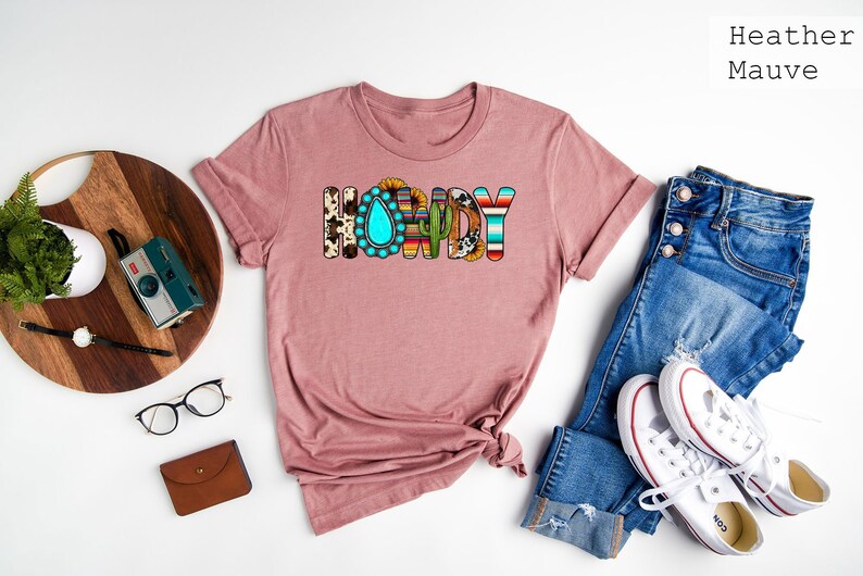 May include: A heather mauve t-shirt with the word "Howdy" printed on the front. The letters are decorated with various southwestern patterns, including turquoise, sunflowers, and cacti.