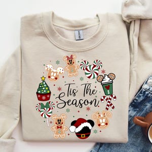 May include: A beige sweatshirt with a Christmas-themed design featuring Mickey Mouse characters, candy canes, and the text "Tis the Season".