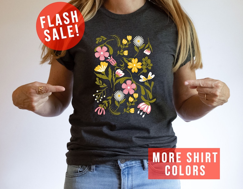 May include: A dark grey t-shirt with a floral print featuring pink, yellow, and green flowers. The text "MORE SHIRT COLORS" is printed in red at the bottom of the image.