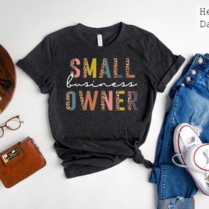 Pode incluir: Camiseta cinza escuro heather com o texto "SMALL business OWNER" em letras coloridas com um padr&atilde;o de leopardo.