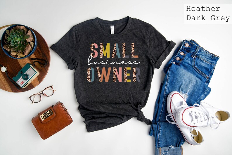 Pode incluir: Camiseta cinza escuro heather com o texto "SMALL business OWNER" em letras coloridas com um padr&atilde;o de leopardo.
