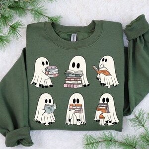 K&ouml;nnte beinhalten: Olivgr&uuml;nes Sweatshirt mit sechs Cartoon-Geistern, die B&uuml;cher halten oder lesen. Die Geister sind wei&szlig; mit schwarzen Augen, und die B&uuml;cher haben verschiedenfarbige Buchr&uuml;cken. Das Sweatshirt hat eine einfarbige gr&uuml;ne Farbe.