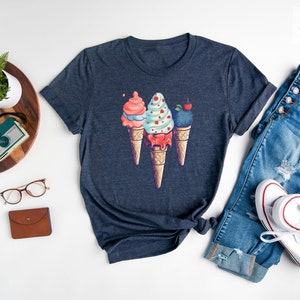 May include: A heather navy blue t-shirt with a graphic of three ice cream cones. The cones are topped with different flavors of ice cream, including strawberry, vanilla, and blueberry. The shirt is laid out on a white surface with a pair of glasses, a watch, a camera, and a wallet.