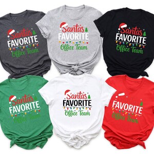 Puede incluir: Seis camisetas de varios colores, incluyendo gris, negro, blanco, verde y rojo. Cada camiseta presenta el texto "Santa's Favorite Office Team" en un diseño festivo con un gorro de Papá Noel y luces navideñas.