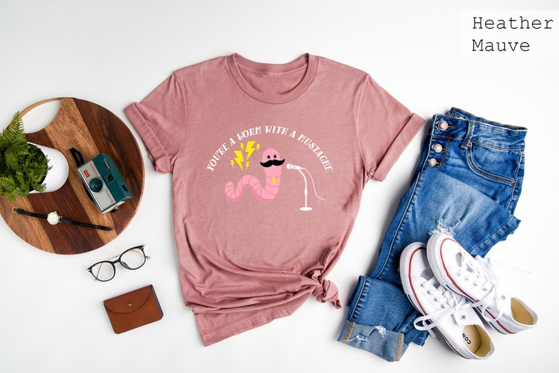 May include: A heather mauve t-shirt with a pink worm wearing a mustache and holding a microphone. The text on the shirt reads "You're a worm with a mustache".