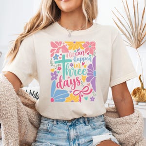 Puede incluir: Camiseta color crema con un diseño floral colorido y el texto "A lot can happen in three days". El diseño incluye una cruz, flores y un lazo. Es una prenda de vestir informal.