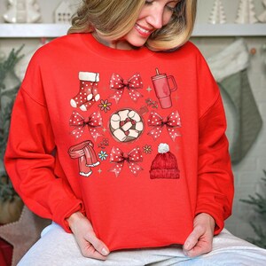 May include: A red sweatshirt featuring a festive design with illustrations of a red beanie, socks, bows, a scarf, a mug, and a cup of hot chocolate with marshmallows. The design includes white polka dots and floral accents, perfect for the holiday season.