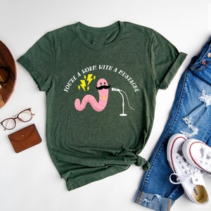 May include: Forest green t-shirt with a pink worm with a mustache holding a microphone. The text on the shirt reads "You're a worm with a mustache".