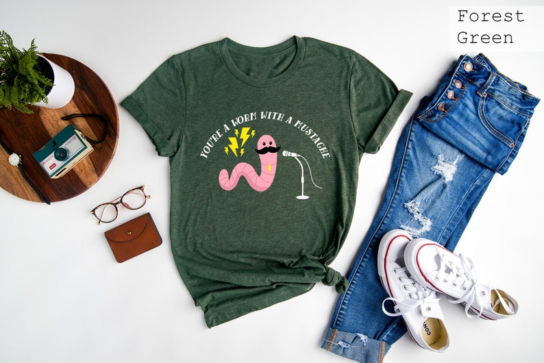 May include: Forest green t-shirt with a pink worm with a mustache holding a microphone. The text on the shirt reads "You're a worm with a mustache".