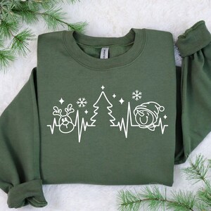 K&ouml;nnte beinhalten: Olivgr&uuml;nes Sweatshirt mit wei&szlig;er Grafik eines Rentiers, Weihnachtsbaums und Schneemanns, verbunden durch eine Herzschlaglinie. Das Design beinhaltet Schneeflocken und Sterne, passend zur Weihnachtszeit.