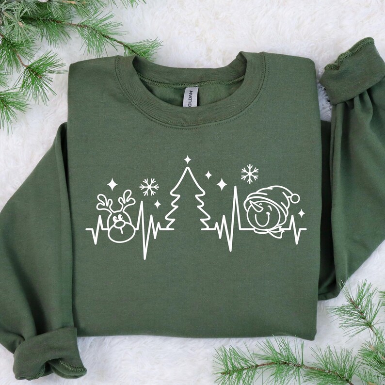 K&ouml;nnte beinhalten: Olivgr&uuml;nes Sweatshirt mit wei&szlig;er Grafik eines Rentiers, Weihnachtsbaums und Schneemanns, verbunden durch eine Herzschlaglinie. Das Design beinhaltet Schneeflocken und Sterne, passend zur Weihnachtszeit.