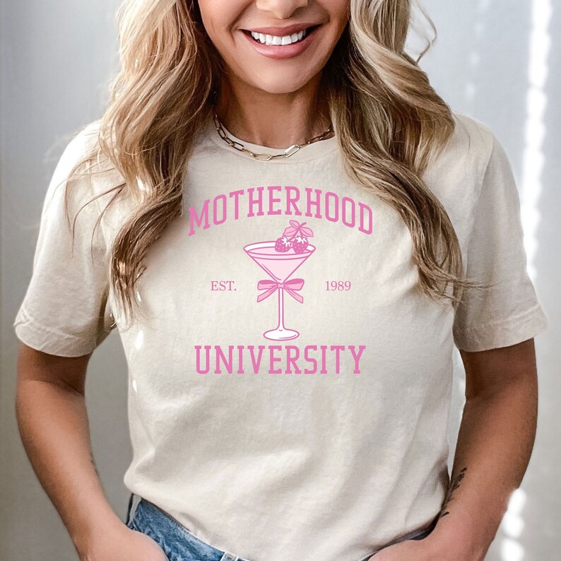 May include: A beige t-shirt with a pink graphic design that says "Motherhood University" with a pink martini glass with strawberries and a pink bow. The text "Est. 1989" is below the martini glass.
