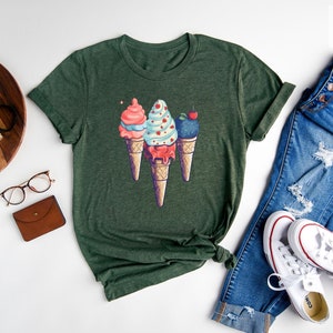 May include: A forest green t-shirt with a graphic of three ice cream cones. The cones are topped with pink, blue, and white ice cream with sprinkles and a cherry.