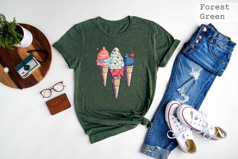 May include: A forest green t-shirt with a graphic of three ice cream cones. The cones are topped with pink, blue, and white ice cream with sprinkles and a cherry.