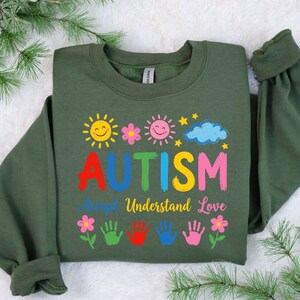 May include: Green sweatshirt featuring the word "AUTISM" in various colours, with the words "Accept, Understand, Love." The design includes smiling suns, flowers, a cloud, stars, and handprints.