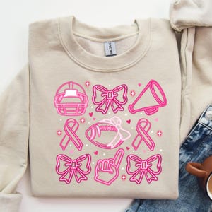May include: A beige sweatshirt with a pink design featuring a football, bows, a ribbon, a megaphone, and a raised index finger. The design is reminiscent of a breast cancer awareness theme.