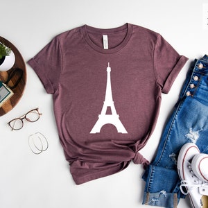 Może przedstawiać: Koszulka w kolorze brązowym melanżowym z białą sylwetką Wieży Eiffla. Koszulka leży na białym tle z okularami, aparatem fotograficznym, rośliną, dżinsami i białymi butami sportowymi.