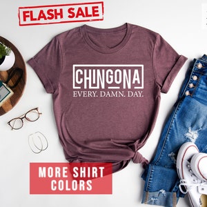 May include: A heather maroon t-shirt with the text "CHINGONA EVERY. DAMN. DAY." printed in white. The shirt is laid flat on a white surface with a pair of jeans, white sneakers, and other accessories.