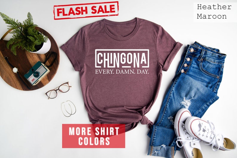 May include: A heather maroon t-shirt with the text "CHINGONA EVERY. DAMN. DAY." printed in white. The shirt is laid flat on a white surface with a pair of jeans, white sneakers, and other accessories.